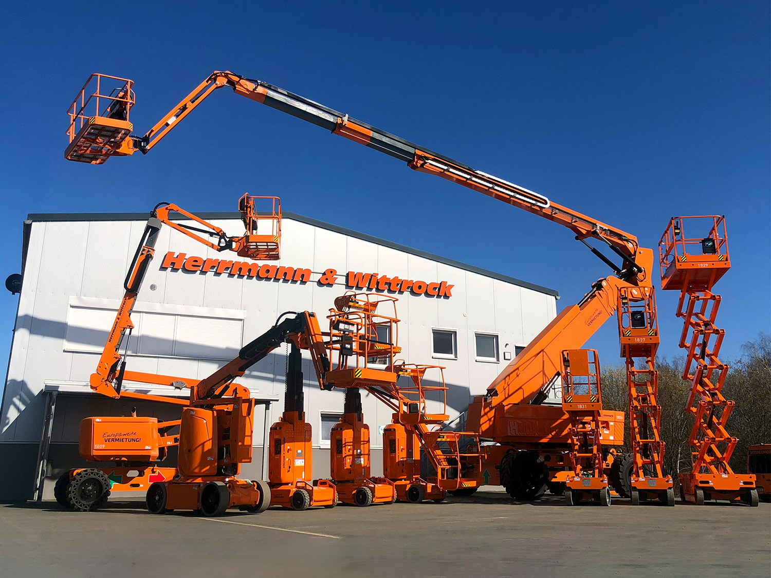 Vor dem Werkstattgebäude von Herrmann & Wittrock ist ein Sortiment an verschiedenen Hebebühnen zu sehen, die orange lackiert sind.