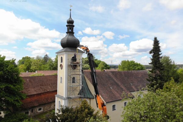 Drohnenbild zeigt den Ladekran von Herrmann & Wittrock vor dem Kirchturm in Thumsenreuth bei Krummennaab. Auf einem Gerüst stehen Mitarbeiter der Firma Bayreuther Turmuhren bereit, um mithilfe des Ladekrans die zwei alten Glocken herauszuheben, die ausgetauscht werden sollen.